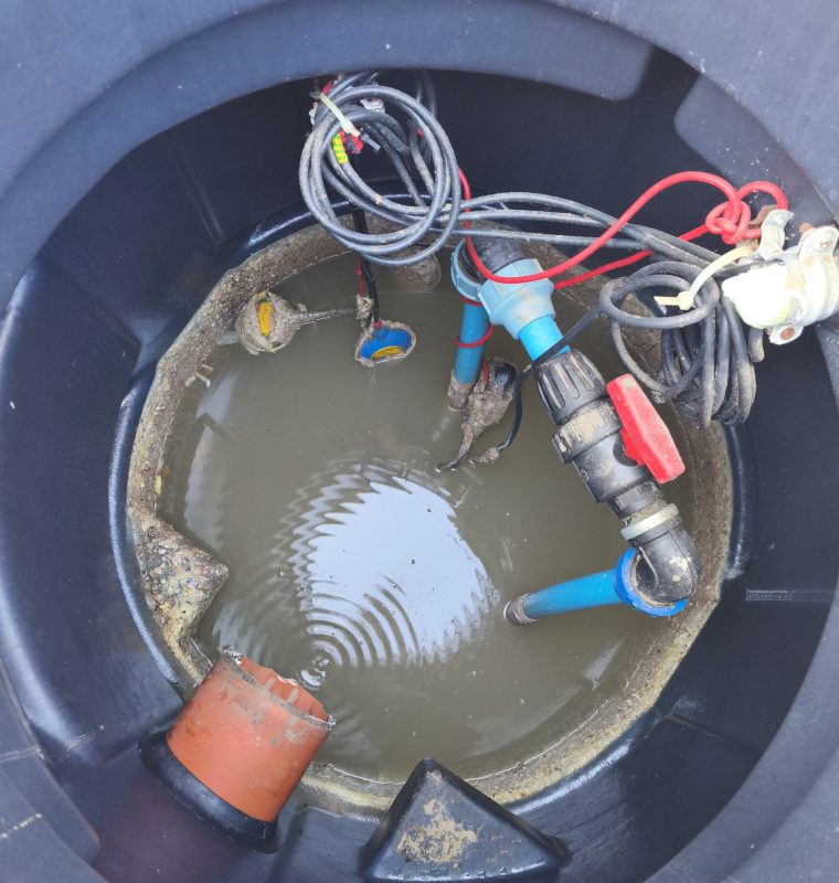 Obraz przedstawia wnętrze studzienki technicznej wypełnionej częściowo wodą, w której znajdują się różne instalacje hydrauliczne i elektryczne, w tym przewody, złącza oraz rury podłączone do systemu pompowego.