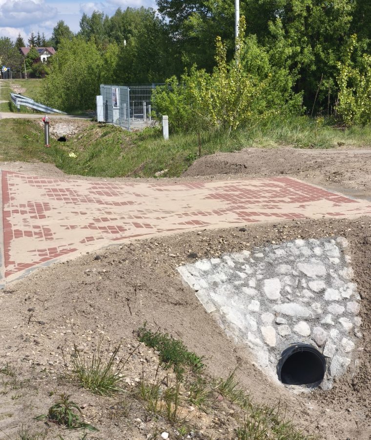 Obraz przedstawia fragment nowo wybudowanego przepustu drogowego z rurą odprowadzającą wodę, wzmocnionego kamieniami i obsypanego ziemią, obok którego znajduje się utwardzona nawierzchnia z kostki brukowej prowadząca przez drogę.