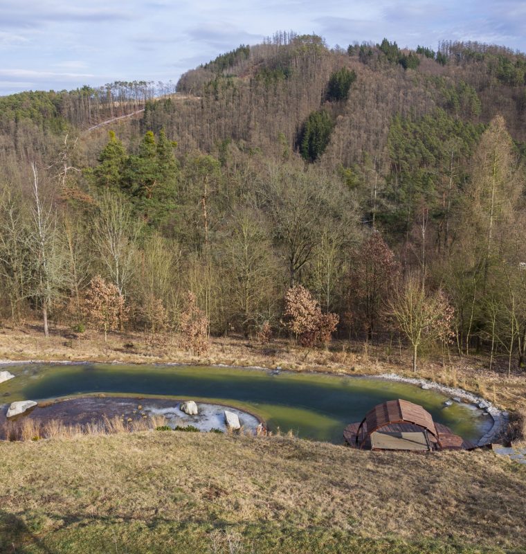 Obraz przedstawia naturalny staw kąpielowy otoczony kamieniami i roślinnością, położony na skraju lasu u podnóża zalesionych wzgórz, z niewielką drewnianą zabudową przy brzegu.