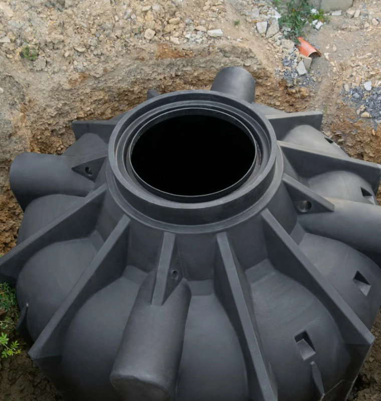Obraz przedstawia dużą plastikową, cylindryczną zbiornikową konstrukcję techniczną umieszczoną w wykopie, która pełni funkcję podziemnego zbiornika – prawdopodobnie na wodę deszczową, ścieki lub jako element systemu kanalizacyjnego.
