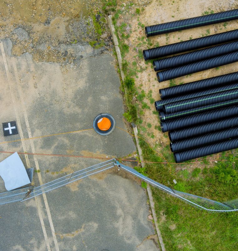 Obraz przedstawia plac budowy sfotografowany z góry, na którym widoczne są składowane czarne rury karbowane przeznaczone do instalacji podziemnych oraz element infrastruktury technicznej w postaci studzienki z pomarańczową pokrywą.